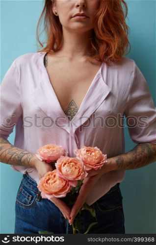 A bouquet of pink roses is holding in hands with tattoo a sexy red-haired girl around a blue background with copy space. Valentine's day concept. Sexy girl with tattoo holding a bouquet of pink roses on a blue background with copy space. Creative layout for postcards
