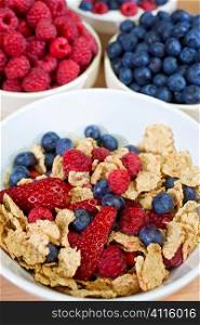 A bowl of healthy breakfast cereals served with strawberries, raspberries and blueberries bowls of which can be seen in the background.