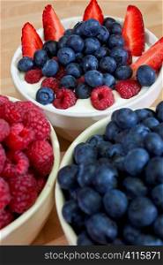 A bowl of healthy breakfast strawberries, raspberries and blueberries in plain yogurt with bowls of raspberries and blueberries in the foreground