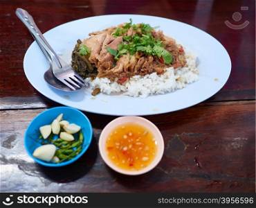 A bowl of rice with pork knuckle meat, served with side dish such as green chili, garlic and sweet chili sauce. Served in a Thai casual restaurant with fork and spoon.