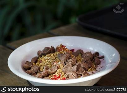 a bowl of strawberry yogurt with granola, fruit and chocolate cornflakes on a wooden table.