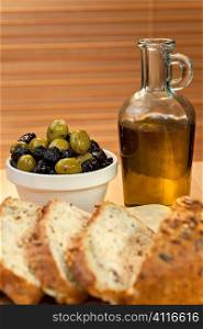 A bowl of stuffed green and black italian olives, a bottle of olive oil and sliced rustic bread in the foreground