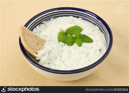 A bowl of tzatziki yoghurt and cucumber dip with a slice of brown farmhouse bread on a woven tablecloth
