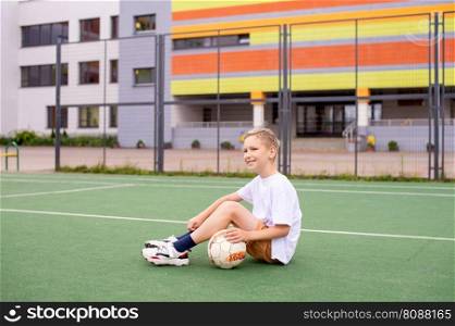 A boy holds a soccer ball while sitting on a soccer field. A teenage boy is sitting on a green field in the school yard with a soccer ball