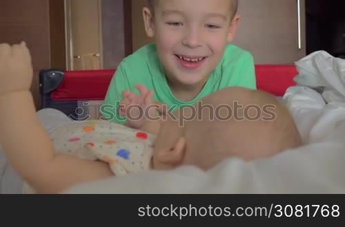 A boy in a green T-shirt is looking at his six months old baby sister who is lying on a bed right in front of him. There is a big happy smile on his face while she is watching her face