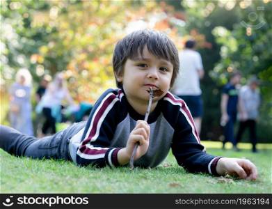 A Boy laying on green grass playing with wooden stick,A Kid holding twig and looking at camera with smiling face with blurry colourful of autumn leaves.Child playing outdoor in the park in fall season