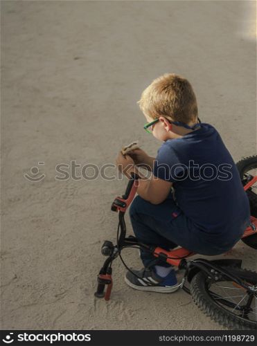 A boy stops playing with his bike to sit on it and eat a sandwich