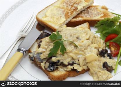 A breakfast or light lunch of scrambled eggs on toast made with fried mushrooms and shallots, served with a sliced cherry tomato and a green salad.