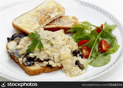 A breakfast or light lunch of scrambled eggs on toast made with fried mushrooms and shallots, served with a sliced cherry tomato and a green salad.