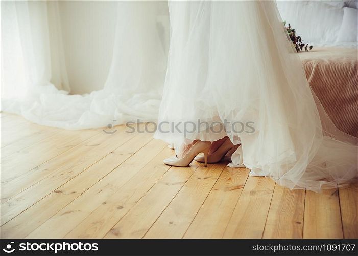A bride in a beautiful wedding white dress and high heeled shoes on wooden floor, next to bed, in preparation for event