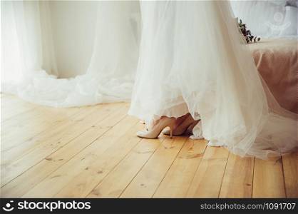 A bride in a beautiful wedding white dress and high heeled shoes on wooden floor, next to bed, in preparation for event