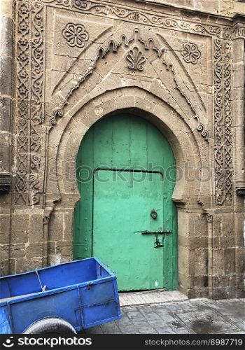 A bright green door contrasts with the blue hand cart and the ancient stone walls of the buildings in the souk of Essaouira, Morocco.