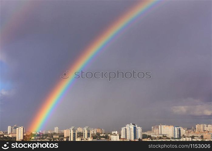 A bright large and dense rainbow in a gray cloudy sky above city houses after the last thunderstorm, image with copy space.. A large bright rainbow in the gray sky above the city after the last thunderstorm.
