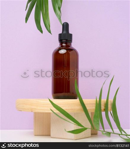A brown glass bottle with a pipette on a wooden podium and green palm leaves. Containers for cosmetics, oils, serums