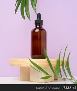 A brown glass bottle with a pipette on a wooden podium and green palm leaves. Containers for cosmetics, oils, serums