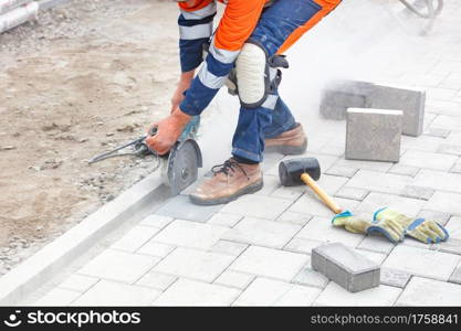 A builder in a blue and orange overalls bends down and cuts off the paving slabs with a grinder for subsequent even laying and leaves behind a cloud of dust. Close-up, selective focus, copy space.. A builder uses a grinder and a diamond cutting disc to cut paving slabs in a cloud of cement dust.