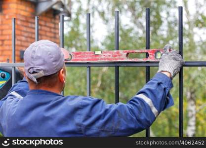 A builder is using an old level to level a metal fence while he is installing a metal fence around a brick house.. The builder uses an old level to level the metal fence when it is being installed.