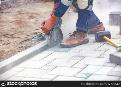 A builder?s hands tensely cut paving slabs in a cloud of cement dust with a grinder and diamond saw blade in front of the workplace. Close-up, selective focus, copy space.. The hands of a builder with the help of a grinder cut the paving slabs against the backdrop of the work site. Close-up, blurred background.