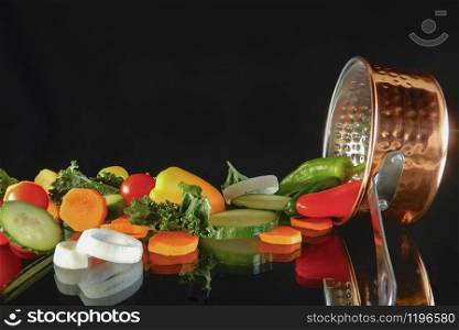 A bunch of assorted freshly sliced vegetables resting next to a saucepan on top of a glass table on a dark background. Healthy and vegan food concept.