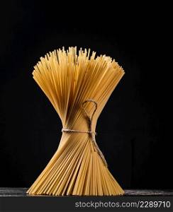 A bundle of spaghetti dry tied with a rope stands on the table. On a black background. High quality photo. A bundle of spaghetti dry tied with a rope stands on the table.