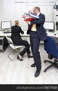 A businessman carrying an armful of dossiers over to his colleague, who&rsquo;s organising her desk