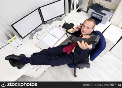 A businessman, comfortable with his feet on his desk, giving the viewer a second call whilst on the phone on another line, in front of a dossier, and a dual monitor displaying financial graphs
