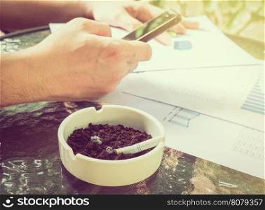 A businessman is seriously working with mobile phone looking at graph paper with cigarette butt in ashtray, selective focus of business and financial working concept.