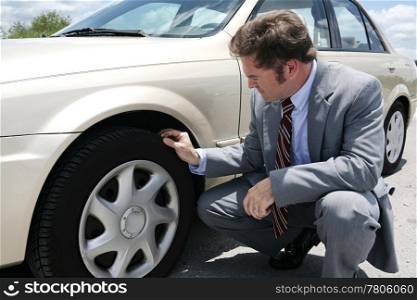 A businessman on the road with a flate tire. He has just discovered the screw that caused the tire to go flat.