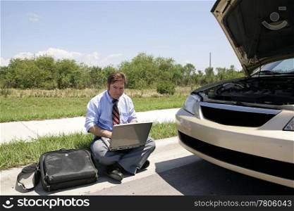 A businessman sitting on the curb working beside his broken down car.