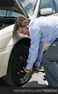 A businessman with a flat tire on the road strains to uncrew the lug nuts.