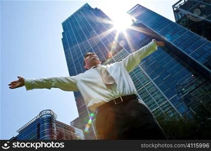 A businessman with his arms out stretched &acute;messiah like&acute; in a modern city environment with the sun bursting over glass fronted office buildings behind him