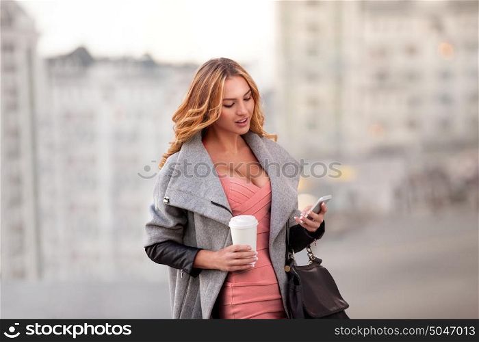 A businesswoman checking email via mobile phone and holding a coffee cup against urban scene.