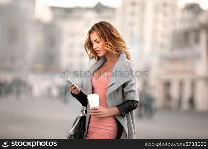 A businesswoman checking email via mobile phone and holding a coffee cup against urban scene.