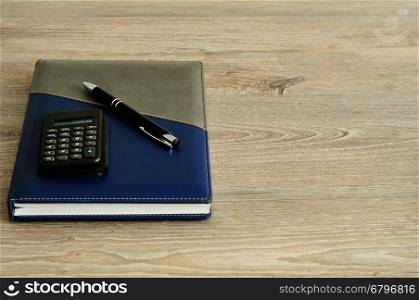 A calculator and pen on top of a note book displayed on a wooden background