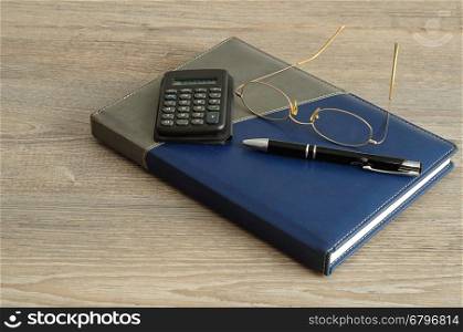 A calculator, pen and reading glasses on top of a note book displayed on a wooden background