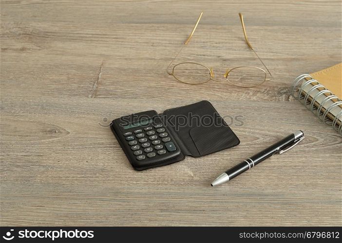 A calculator, pen, reading glasses and notebook displayed on a wooden background