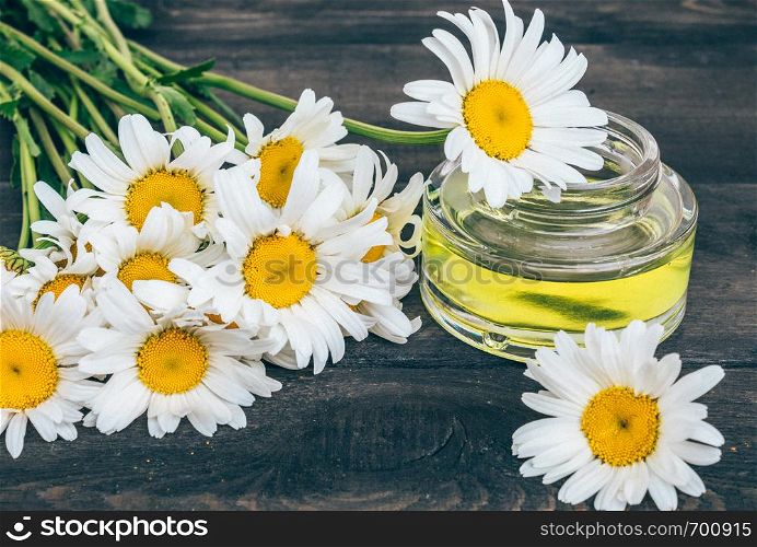 A can of light essential oil from chamomiles stands on dark boards near chamomile flowers.. On the boards near the chamomile flowers there is a bank of light chamomile essential oil.