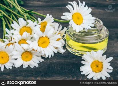 A can of light essential oil from chamomiles stands on dark boards near chamomile flowers.. On the boards near the chamomile flowers there is a bank of light chamomile essential oil.
