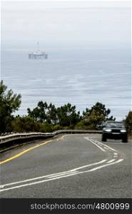A car drives up a steep mountain pass with an oil drill platform in the ocean in the background