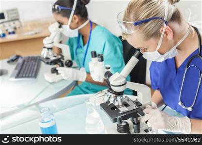 A caucasian female medical or scientific researcher or doctor using her microscope in a laboratory with her Asian Indian colleague beside her.