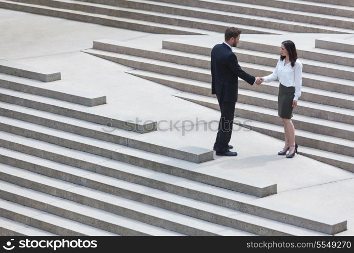 A caucasian man businessman and an Asian woman businesswoman shaking hands on a deal standing on modern city steps