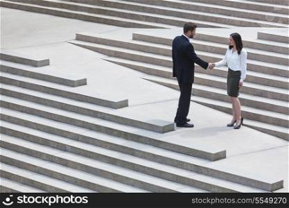 A caucasian man businessman and an Asian woman businesswoman shaking hands on a deal standing on modern city steps