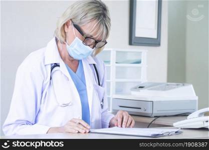 A caucasian senior female doctor wearing mask writing patient&rsquo;s application for registering at reception in hospital