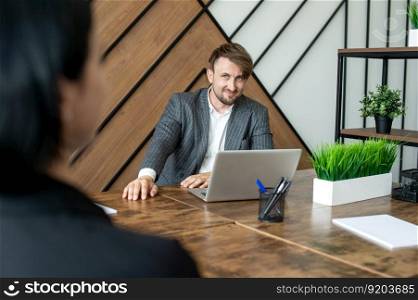 A cheerful man in a jacket is sitting at his desk in the office with a laptop. Has a conversation with a colleague. The concept of the problem of procrastination in the workplace. Selective focus. A cheerful man in a jacket is sitting at his desk in the office with a laptop.
