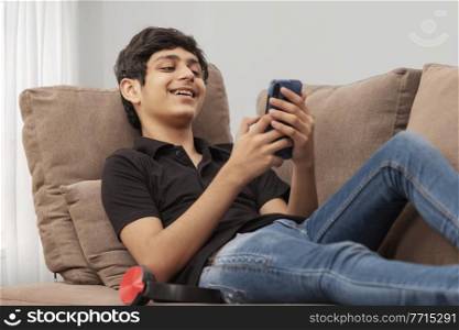 A CHEERFUL TEENAGER RESTING ON SOFA AND USING MOBILE PHONE