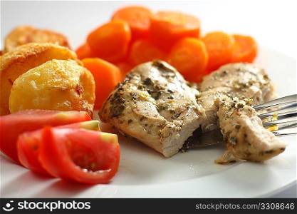 A chicken breast, baked with herbs, being cut. The meal also features carrots, roast potatoes and sliced tomato
