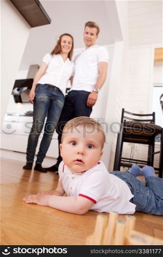 A child on the floor looking intently at the camera, parents in the background