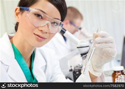 A Chinese Asian Oriential female medical or scientific researcher or doctor using looking at a test tube of clear liquid in a laboratory, with her colleague out of focus behind her.