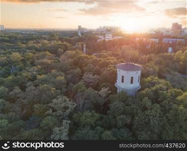 A city in the distance and a green park with a tower and people on the roof against the sky with the sunset. Aerial view from the drone. Aerial view of the drone on the green park with a round tower and the city in the distance at sunset