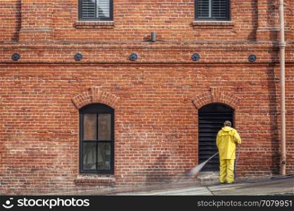 a city worker in yellow rain coat wahsing sidewalk with a hose in front of brick house